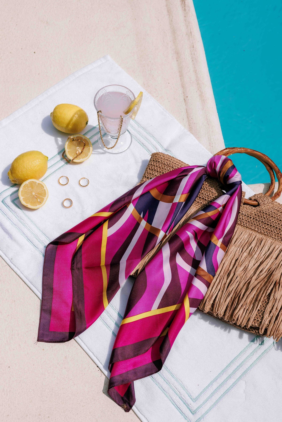 A patterned silk scarf with bold magenta, purple, white, and yellow stripes is styled on a white beach towel next to a woven straw bag, lemons, and a drink by a pool.