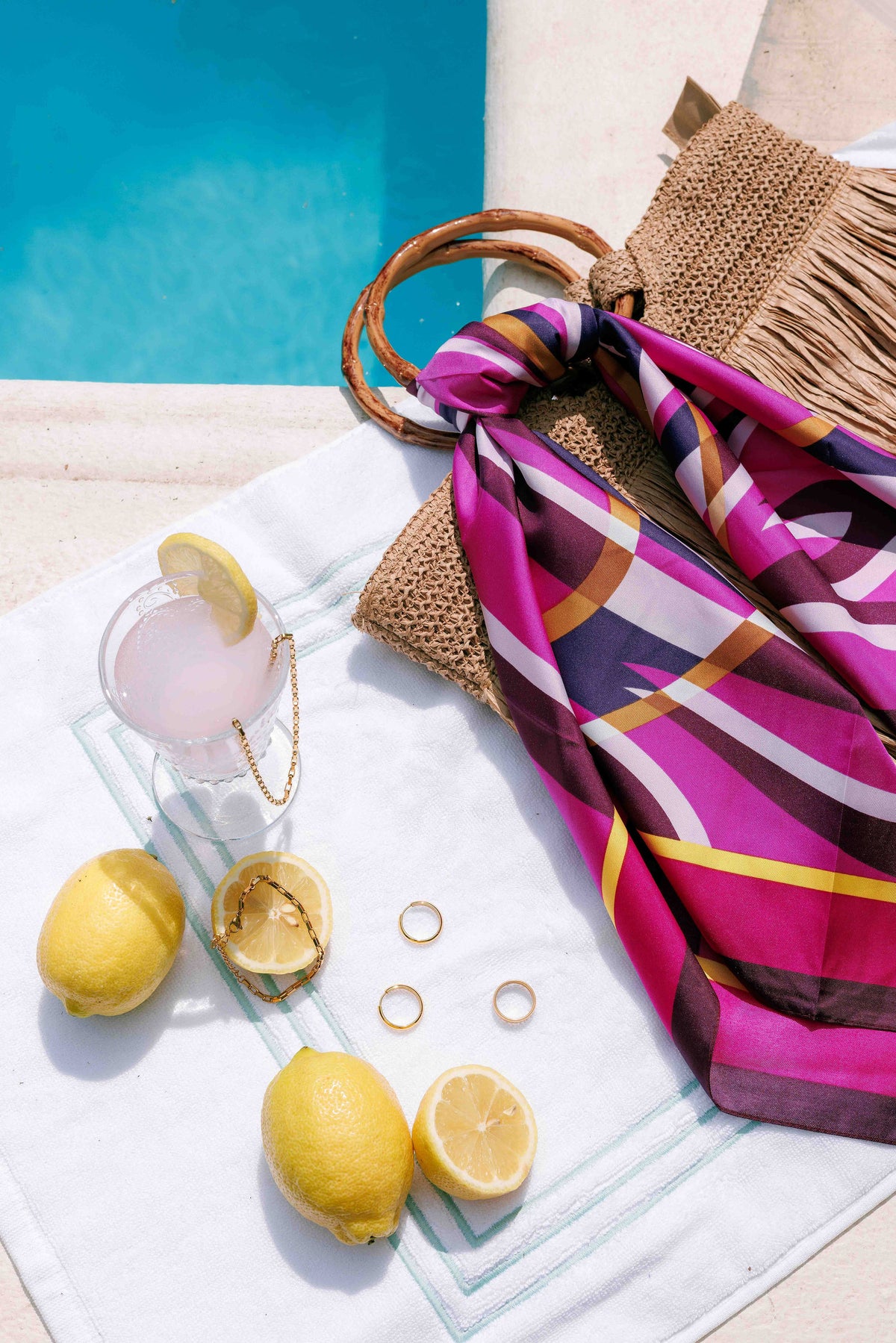 A styled poolside scene with a bright pink, purple, white, and gold striped scarf draped over a woven straw bag, alongside lemons, a drink, rings, and a blue swimming pool in the background.