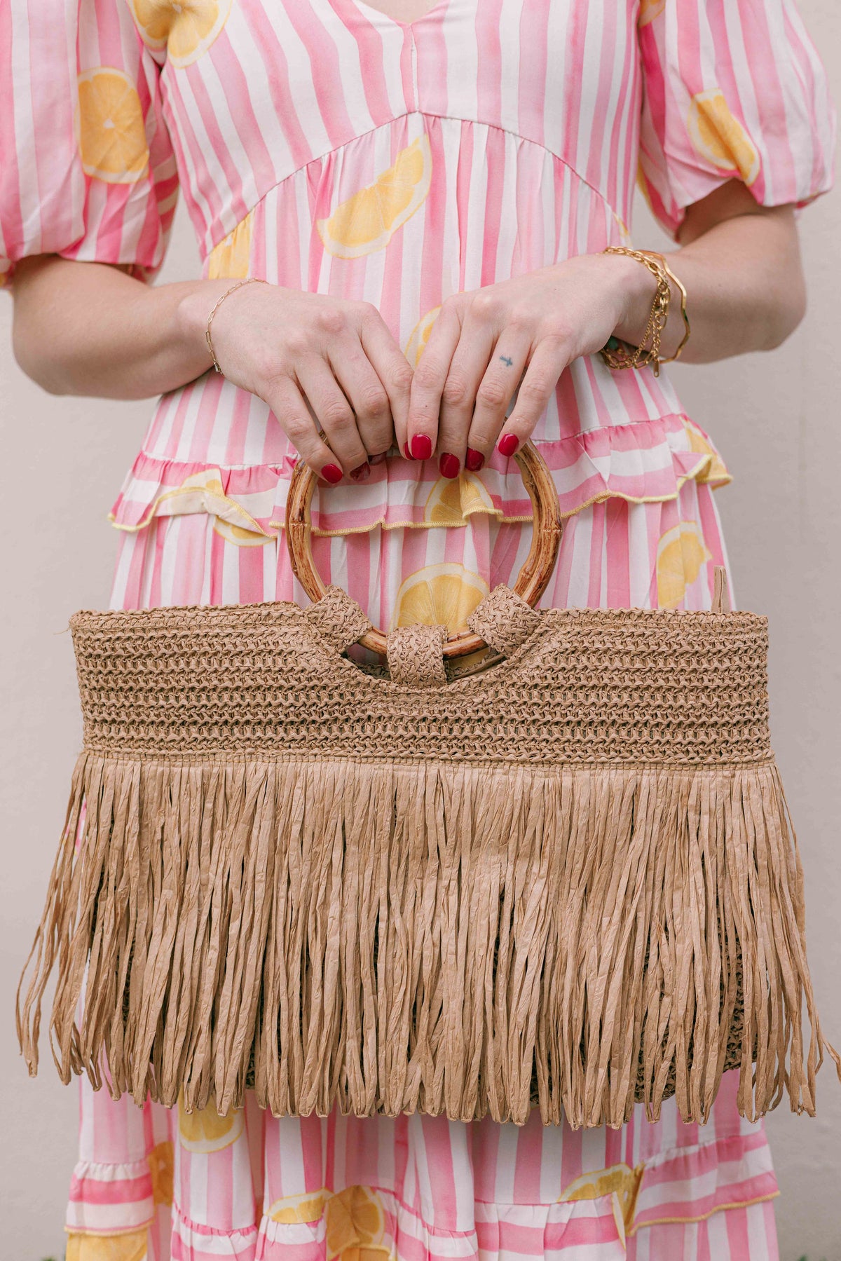 A woman in a pink and white striped dress with yellow citrus prints holds a large woven straw handbag with a fringe bottom and round wooden handles.