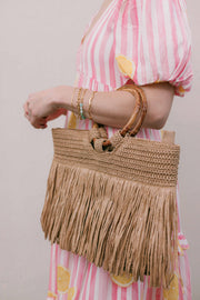 A woman wearing a pink and white striped dress with lemon prints holds a natural straw fringe handbag with wooden handles against a light beige wall.